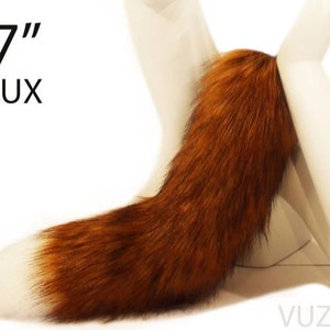 Puede incluir: Una cola de piel sintética de 43 cm de largo, de color marrón rojizo. La cola está unida a una superficie blanca.