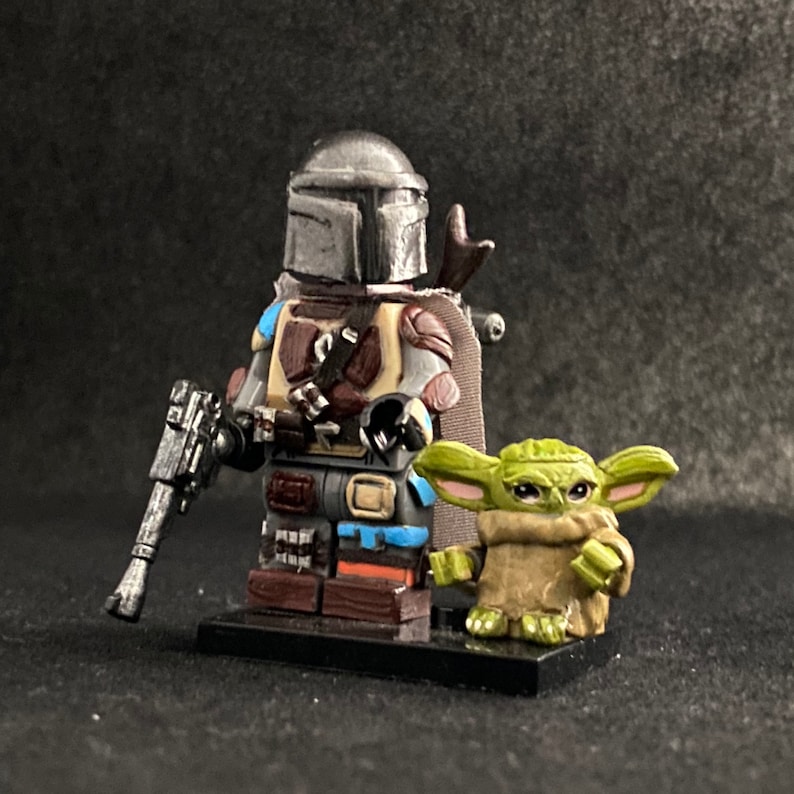 May include: A custom-made LEGO minifigure of the Mandalorian from the Star Wars series, wearing his signature helmet and armor. He is holding a blaster rifle and standing next to a small green alien figure, Grogu, also known as Baby Yoda.