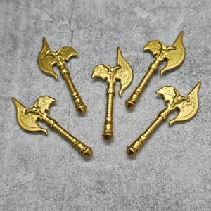 May include: Five gold-colored axe-shaped toy weapons with intricate details. The axes have a curved blade and a handle with a decorative design.
