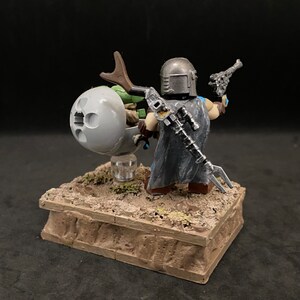 May include: A custom-painted LEGO minifigure of The Mandalorian from the Star Wars series, holding a blaster pistol and riding a speeder bike with a small green alien figure. The minifigure is wearing a silver helmet and a grey cloak. The speeder bike is grey and has a brown handle. The minifigure and speeder bike are on a brown base.