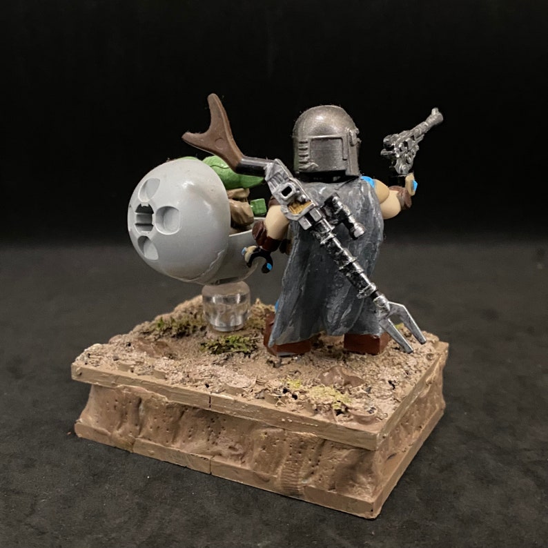 May include: A custom-painted LEGO minifigure of The Mandalorian from the Star Wars series, holding a blaster pistol and riding a speeder bike with a small green alien figure. The minifigure is wearing a silver helmet and a grey cloak. The speeder bike is grey and has a brown handle. The minifigure and speeder bike are on a brown base.