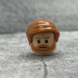 May include: A brown Lego minifigure head with a brown hair piece and a facial expression of concern. The minifigure has a light brown beard and mustache.