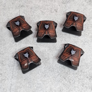 Könnte beinhalten: Fünf braune Spielzeug-Rüstungsstücke aus Kunststoff mit einem Wolfs-Emblem auf der Vorderseite. Jedes Stück hat eine schwarze Basis und eine braune, strukturierte Oberfläche.