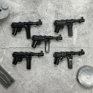 May include: Five black plastic toy machine guns. The guns are all the same design, with a long barrel, a magazine, and a grip. They are all lying on a grey fabric surface.
