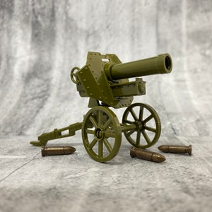 May include: A green toy cannon with wheels and a barrel pointing to the right. There are three brown toy cannonballs in front of the cannon.