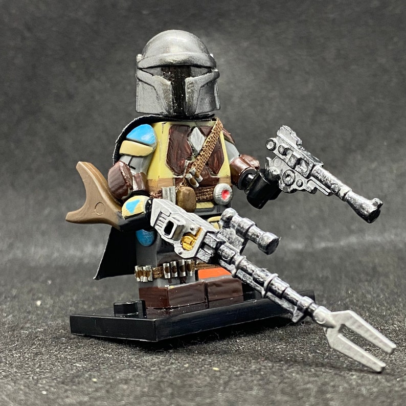 May include: A custom-painted Lego minifigure of a Mandalorian with a blue and brown helmet, holding a blaster rifle and a pistol. The figure is wearing a brown leather vest and a blue and brown cape.
