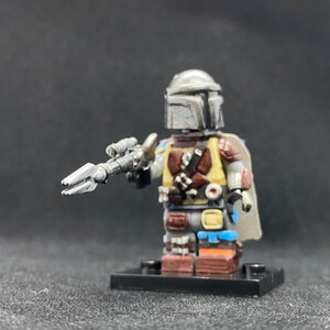 May include: A custom-made Lego minifigure of a Mandalorian character. The figure is wearing a silver helmet with a weathered look, a brown and yellow vest, and a blue and brown cape. The figure is holding a silver blaster rifle.