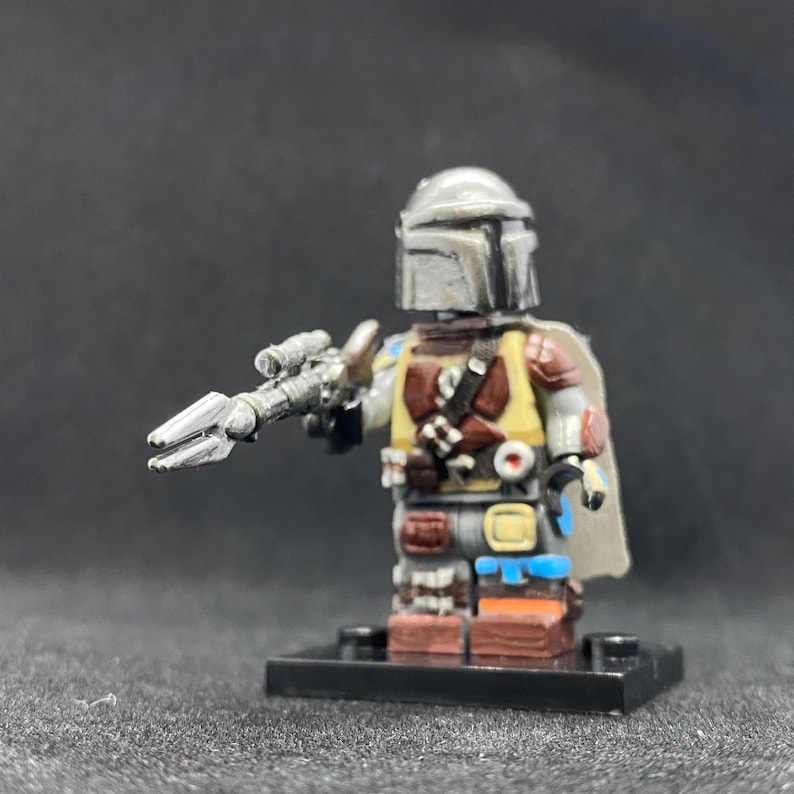 May include: A custom-made Lego minifigure of a Mandalorian character. The figure is wearing a silver helmet with a weathered look, a brown and yellow vest, and a blue and brown cape. The figure is holding a silver blaster rifle.