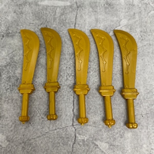 May include: Five gold-colored plastic swords with a curved blade and a decorative design. The swords are all the same size and shape.