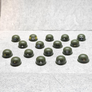 May include: A collection of olive green miniature military helmets. Each helmet features different insignia, including crosses, stars, and chevrons. The helmets are arranged on a light gray surface.