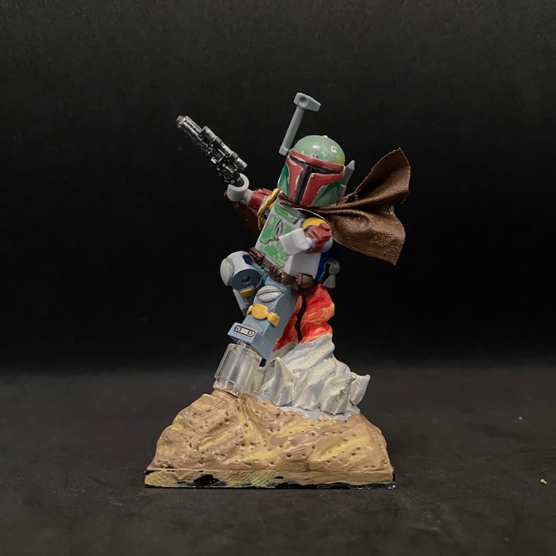 May include: A custom-painted Lego minifigure of Boba Fett, standing on a brown and gray base. The figure is holding a blaster and has a brown cape.