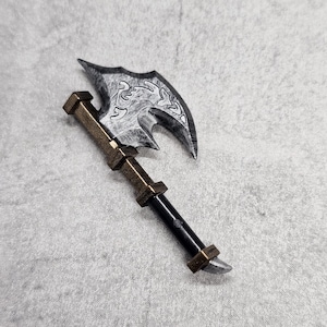 May include: A miniature battle axe with a silver blade and a bronze handle. The blade has a decorative design. The handle is black with bronze accents. The axe is a fantasy-themed item.
