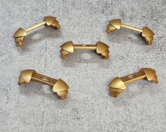 Puede incluir: Cinco piezas decorativas de metal de color dorado, con forma curva y un pequeño agujero en el centro. Las piezas están dispuestas en forma circular sobre una superficie gris texturizada.