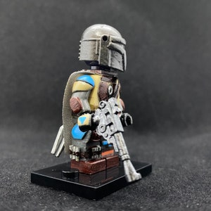 May include: A custom-painted Lego minifigure of a Mandalorian with a weathered silver helmet, blue and yellow armor, and a silver blaster rifle.