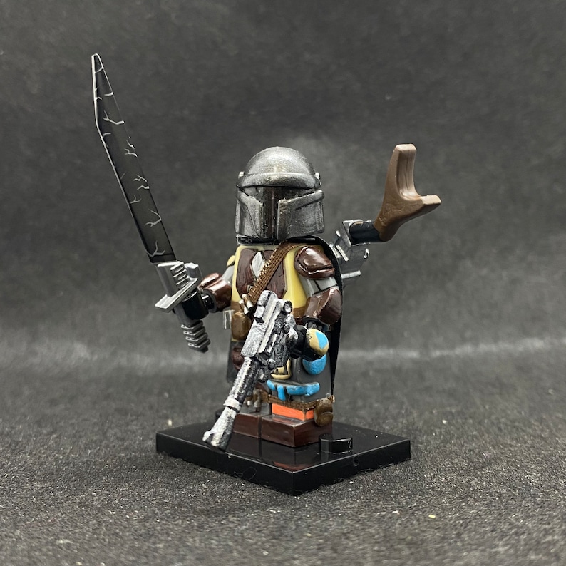 May include: A custom-painted Lego minifigure of a Mandalorian character. The figure is wearing a helmet with a weathered paint job, a brown and tan vest, and a blue and gray cape. The figure is holding a blaster rifle and a silver lightsaber.