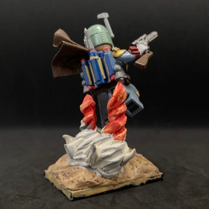 May include: A custom-made Lego minifigure of Boba Fett standing on a rocky base. The figure is wearing a brown cape and has a blue and yellow jetpack. The figure is holding a blaster rifle in one hand and a blaster pistol in the other.