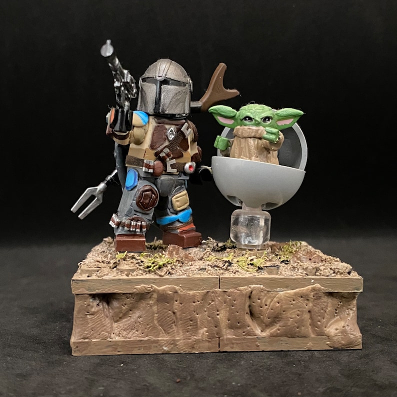 May include: A custom-made LEGO minifigure of The Mandalorian with a blaster rifle, standing next to a LEGO minifigure of Grogu in a floating pod. Both figures are on a brown base.
