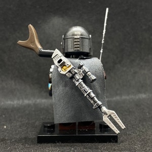May include: A black and silver Lego minifigure of a Mandalorian character with a cape and a blaster rifle. The minifigure is standing on a black base.
