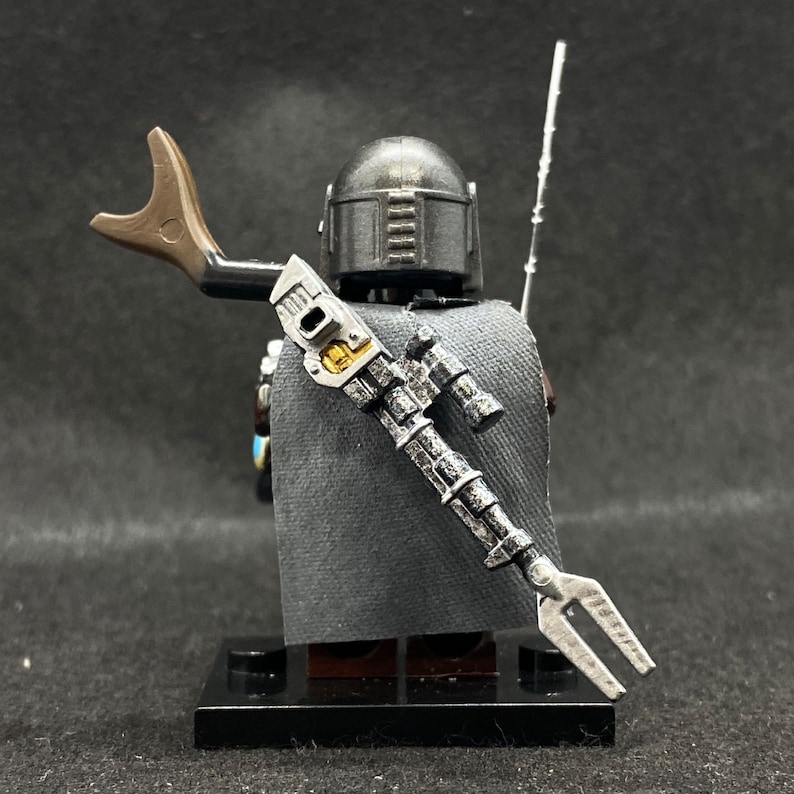May include: A black and silver Lego minifigure of a Mandalorian character with a cape and a blaster rifle. The minifigure is standing on a black base.