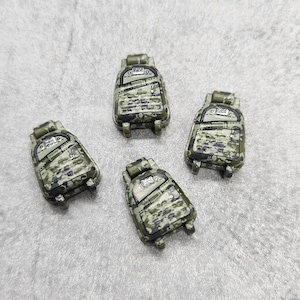 以下が含まれることがあります： 黒いストラップとジッパーが付いた、迷彩柄のミニチュアバックパックが4つ。バックパックは質感のある素材で作られており、リアルな迷彩柄になっています。