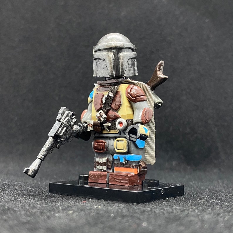 May include: A custom-painted Lego minifigure of the Mandalorian, wearing his iconic helmet and armor. The figure is holding a blaster rifle and has a cape and a jetpack. The figure is standing on a black Lego baseplate.