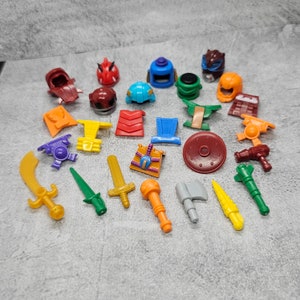 May include: A collection of colourful plastic toy accessories, including helmets, shields, swords, and other fantasy-themed items.