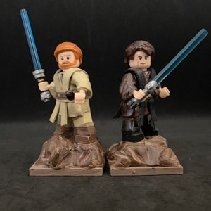 Puede incluir: Dos minifiguras Lego, una con una túnica marrón y un sable de luz azul, la otra con una túnica negra y un sable de luz azul, de pie sobre bases marrones.