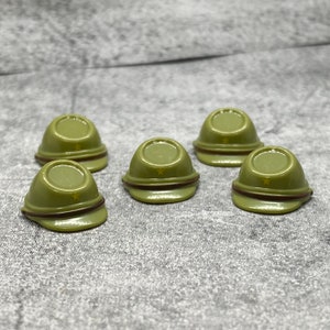 Könnte beinhalten: Fünf grüne Militärhelme im Miniaturformat mit einem gelben Stern auf der Oberseite. Die Helme sind in einem Halbkreis auf einer grauen, strukturierten Oberfläche angeordnet.