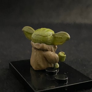 May include: A small green and brown figurine of a character from the Star Wars franchise, sitting on a black base.