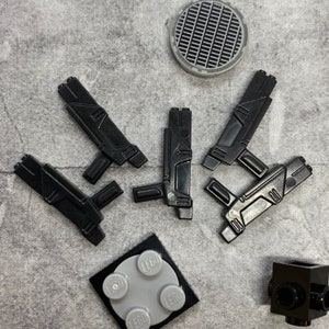 Puede incluir: Cinco pistolas de juguete de plástico negro sobre un fondo de tela gris. También hay cuatro ladrillos LEGO grises con la palabra "LEGO" impresa en ellos.
