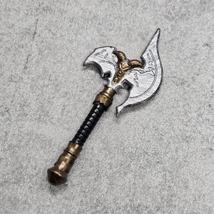 以下が含まれることがあります： 黒の柄が付いた銀と金の斧。斧の頭は、中央に角のある頭蓋骨を持つ詳細なデザインです。