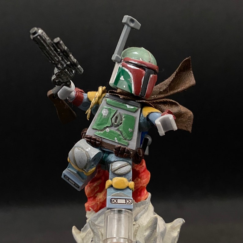 May include: A green and red Lego minifigure of Boba Fett, wearing a brown cape and holding a blaster. The minifigure is standing on a white base.