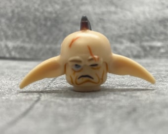 Puede incluir: Una figurita de goblin beige con un sombrero puntiagudo, orejas grandes y una cara detallada. El goblin tiene el ceño fruncido, un ojo cerrado y una mueca.