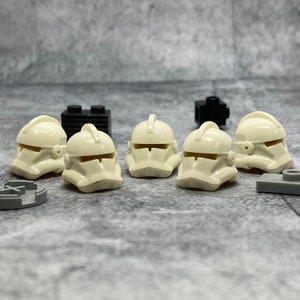 May include: Six white plastic Lego minifigure helmets with a raised design on the front. The helmets are arranged in a row on a gray surface.
