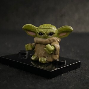 May include: A small green and brown figurine of a character from the Star Wars franchise, known as "The Child" or "Baby Yoda". The figurine is standing on a black base.