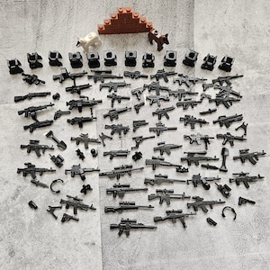 以下が含まれることがあります： 黒いプラスチック製の玩具銃のコレクションで、ライフル、ピストル、マシンガンなどがあります。銃は灰色の表面に並べて置かれています。背景には、小さなプラスチック製のフィギュアとレンガの壁もあります。