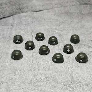 May include: A collection of ten miniature green helmets with different white symbols on top. The symbols include stars, a cross, a heart, and a letter H.