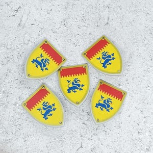 May include: Five yellow shield-shaped emblems with a red top border and a blue lion design. The emblems are arranged on a light gray textured surface. Each shield has a small hole at the top.