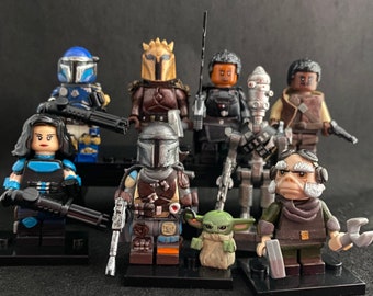 Puede incluir: Una colección de minifiguras de Star Wars, que incluye un Mandaloriano azul y plateado, un Mandaloriano marrón y dorado, un Stormtrooper negro y plateado, un cazarrecompensas marrón y negro, un extraterrestre verde y marrón y un personaje marrón y negro con barba blanca.