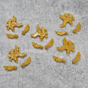 May include: Gold-coloured griffin charms and detached wings scattered on a grey surface. The griffin charms have detailed wings and a stylised body. The wings are separate pieces, also in gold. These charms are suitable for jewellery making or craft projects.