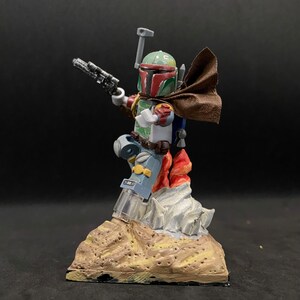 May include: A custom-painted Lego minifigure of Boba Fett, standing on a base with flames and a rocky surface. The figure is wearing a brown cape and holding a blaster.