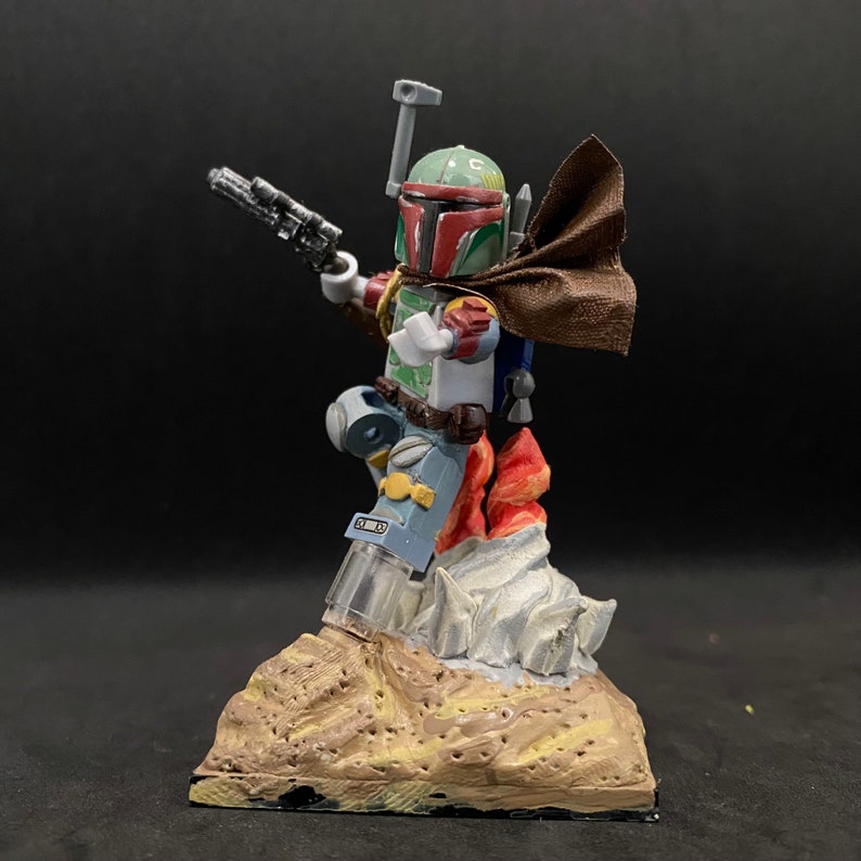 May include: A custom-painted Lego minifigure of Boba Fett, standing on a base with flames and a rocky surface. The figure is wearing a brown cape and holding a blaster.