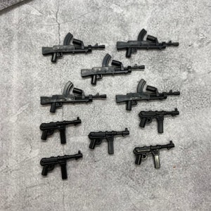 May include: A collection of ten black plastic toy guns, including rifles and pistols. The guns are arranged in a row on a grey textured surface.