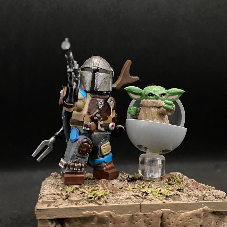 May include: A custom-made Lego minifigure of the Mandalorian, wearing a grey and brown helmet and armor, holding a blaster rifle. The Mandalorian is standing on a brown base with a small green Lego minifigure of Grogu, also known as Baby Yoda, sitting in a floating pod.