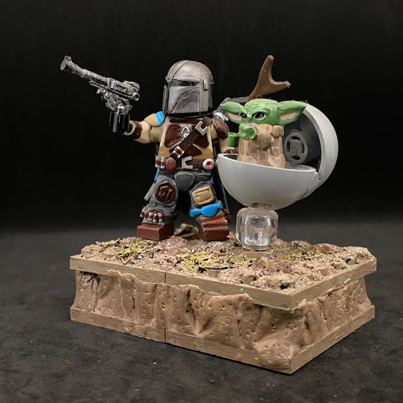 May include: A custom-made LEGO minifigure of the Mandalorian from Star Wars, holding a blaster rifle. He is standing next to a green alien character, known as Grogu, in a floating pod. The figures are on a brown, textured base.