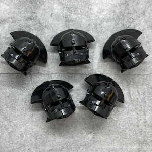 May include: Five black plastic helmets with a face design and a raised ridge on the top. The helmets are arranged in a semi-circle on a gray textured surface.