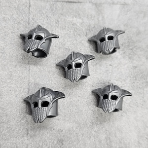 May include: Five silver-coloured helmets with a detailed design. The helmets have a pointed top and a face mask with a grid pattern.