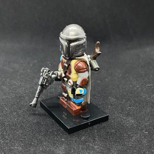 May include: A custom-painted LEGO minifigure of the Mandalorian, wearing his iconic helmet and armor. The figure is holding a blaster rifle and has a cape. The figure is standing on a black base.
