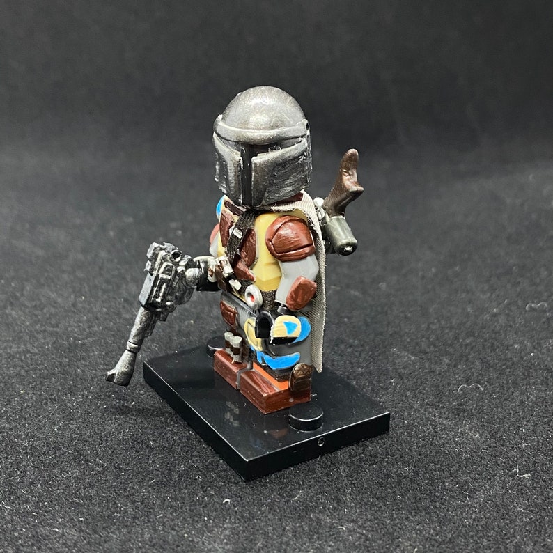 May include: A custom-painted LEGO minifigure of the Mandalorian, wearing his iconic helmet and armor. The figure is holding a blaster rifle and has a cape. The figure is standing on a black base.