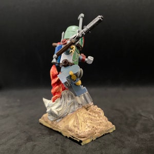May include: A green and brown action figure of Boba Fett standing on a sand-colored base with a red and white flame effect. The figure is holding a blaster rifle in its right hand.